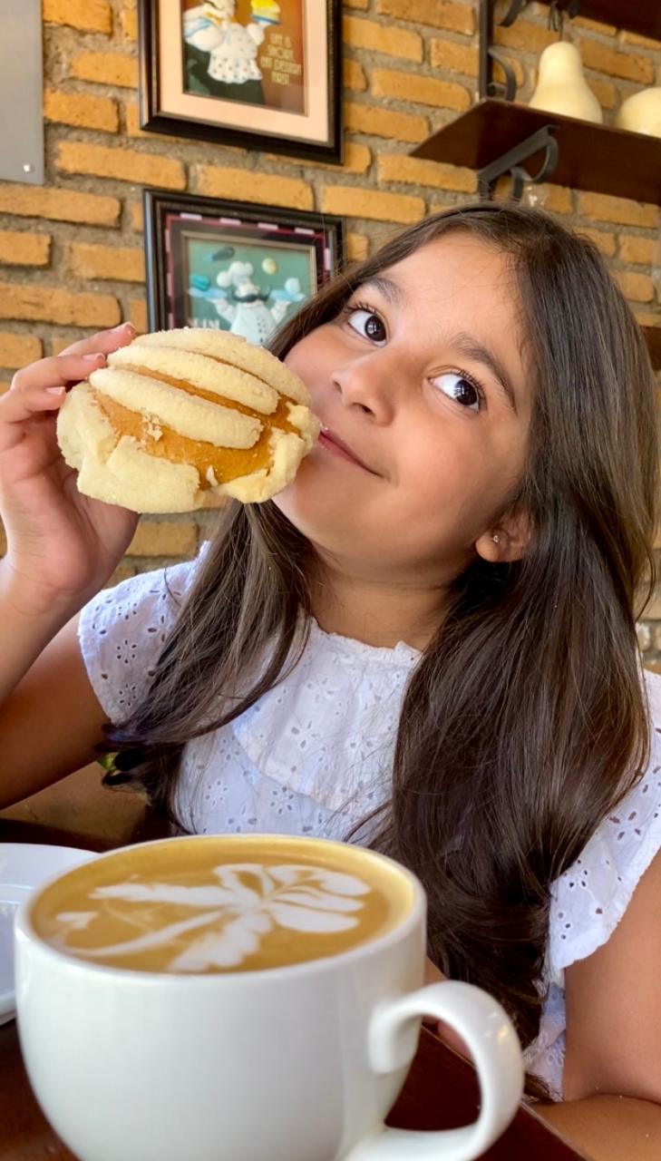 Niña disfrutando un pan dulce junto a un café con arte latte en Pistrina